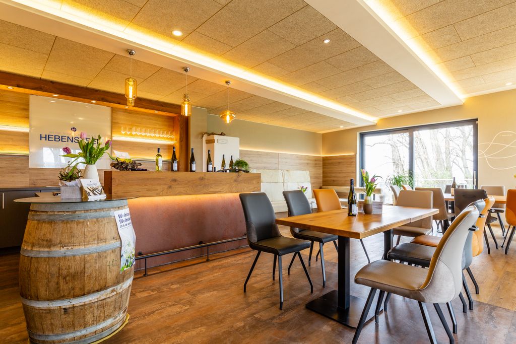 Moderner Verkostungsraum mit Holztischen, Stühlen und einer Bar. Weinflaschen und Blumen dekorieren den Raum.