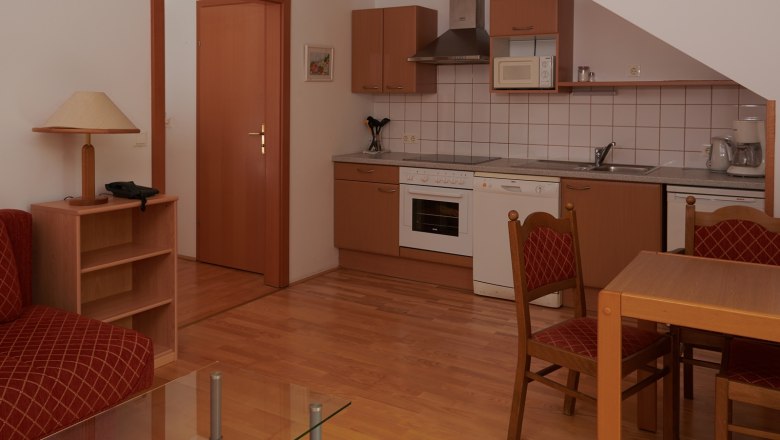 Kompakte K&uuml;che mit Holzschr&auml;nken, Esstisch und St&uuml;hlen, Herd, Sp&uuml;le und Mikrowelle.