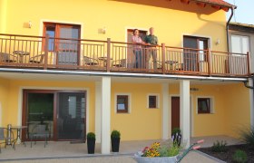 Zweistöckiges gelbes Gebäude mit Balkon, auf dem zwei Personen stehen. Im Vordergrund eine Schubkarre mit Blumen.