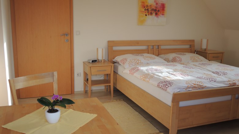 Ein G&auml;stezimmer mit einem Doppelbett, zwei Nachttischen, einem Tisch mit Stuhl und einer Topfpflanze.