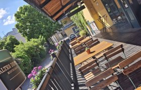 Eine sonnige Terrasse mit Holztischen und -stühlen, umgeben von Pflanzen und einem Gebäude mit gelber Fassade.