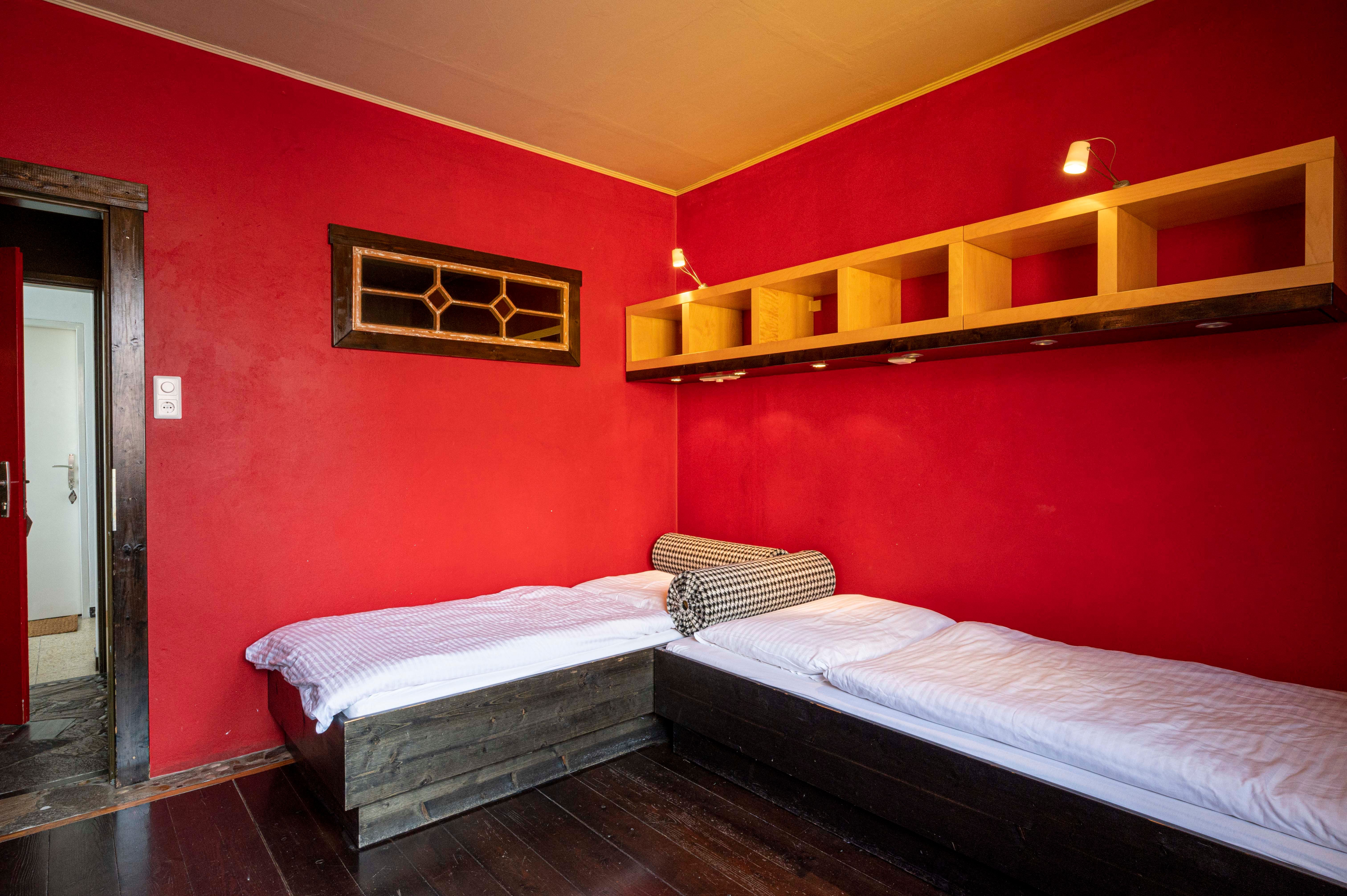 Ein Zimmer mit roten Wänden, zwei Einzelbetten und einem Regal an der Wand.