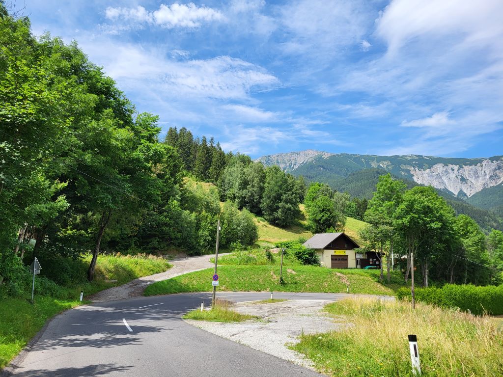 Landschaft mit Straße, Haus und Bergen im Hintergrund.