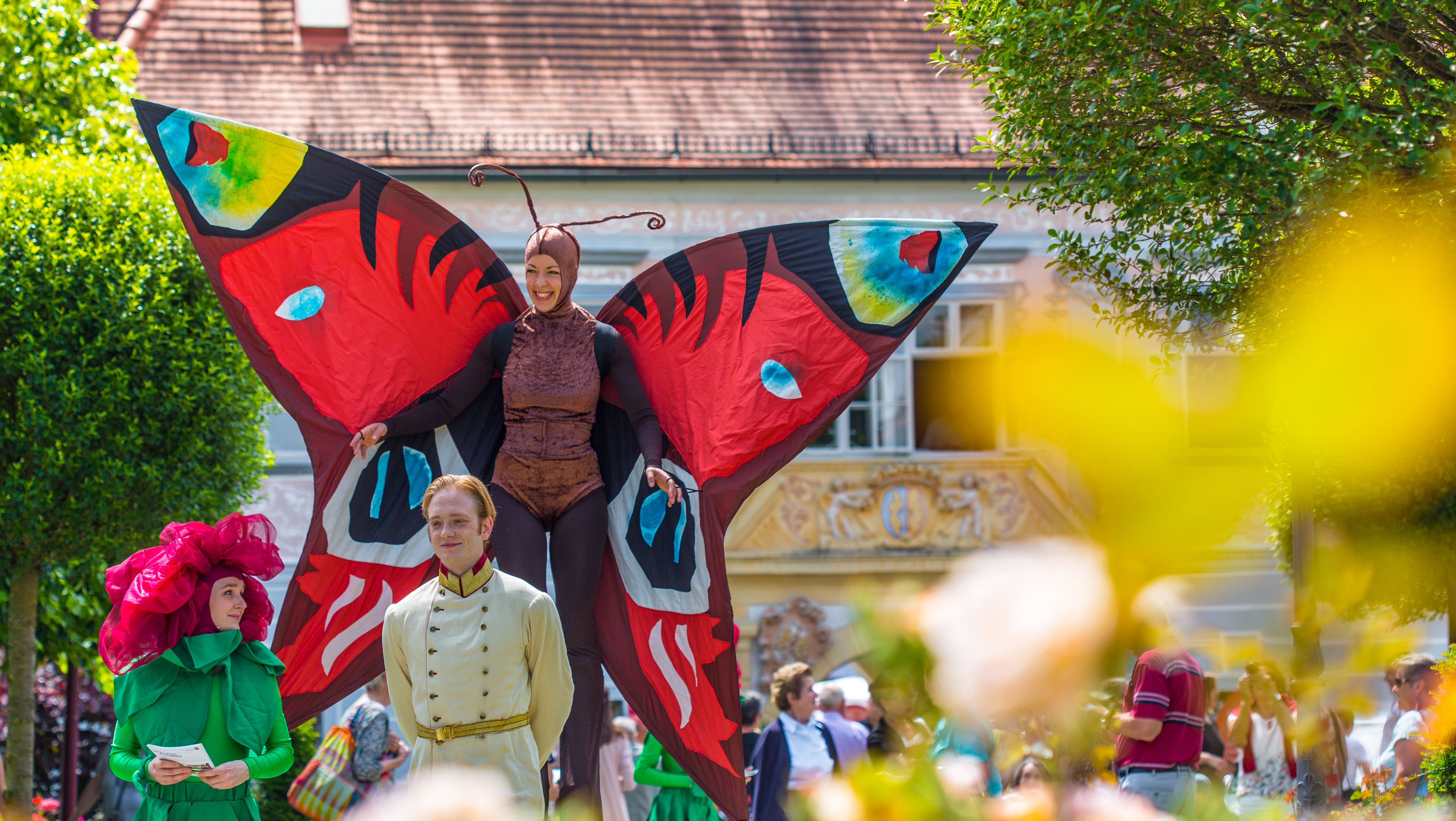 Bunt verkleidete Menschen beim Rosenfest, eine Frau ist auf Stelzen als großer bunter Schmetterling verkleidet