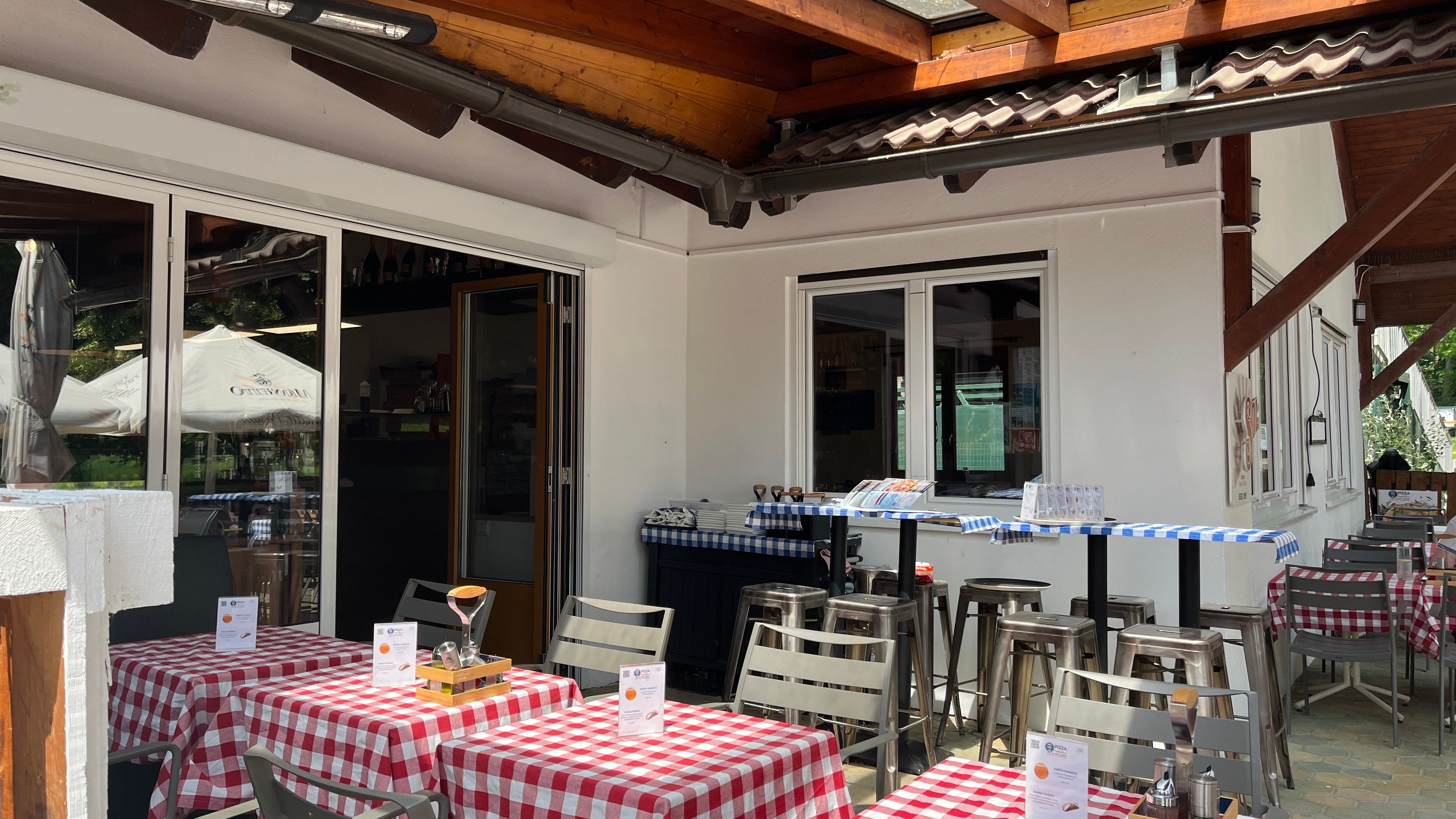 Eingangsbereich der Pizzeria mit Gastgarten. Im Vordergrund sind gedeckte Tische mit roter Tischdecke.