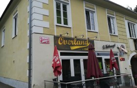 Gelbes Gebäude mit dem Schriftzug 'Overland' und roten Sonnenschirmen.