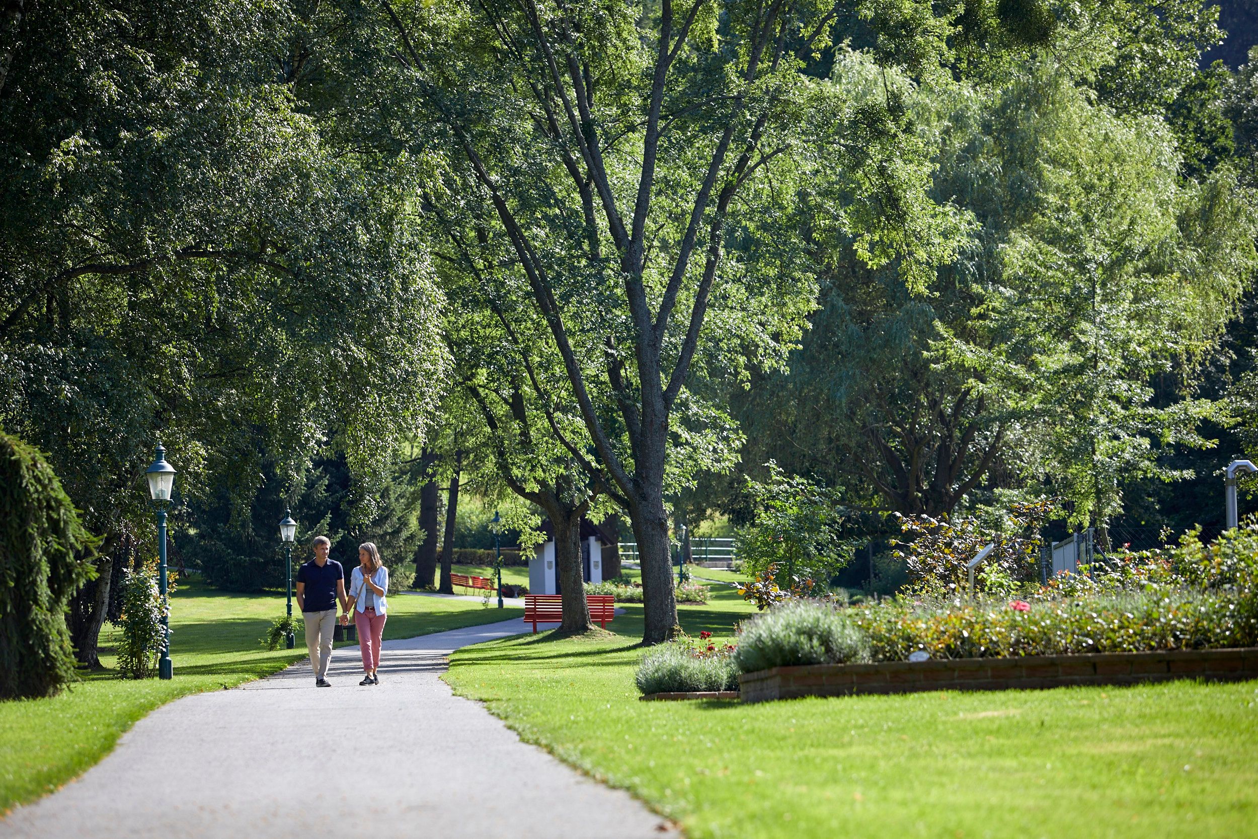 Ein Paar spaziert auf einem Weg im Kurpark Bad Schönau, umgeben von grünen Bäumen und gepflegten Blumenbeeten.