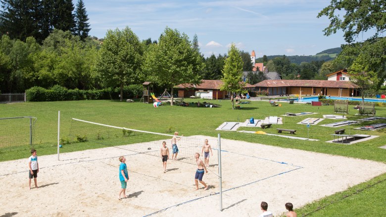 Kinder spielen Volleyball auf einem Sandplatz im Freibad Aspang, umgeben von gr&uuml;ner Wiese und B&auml;umen.
