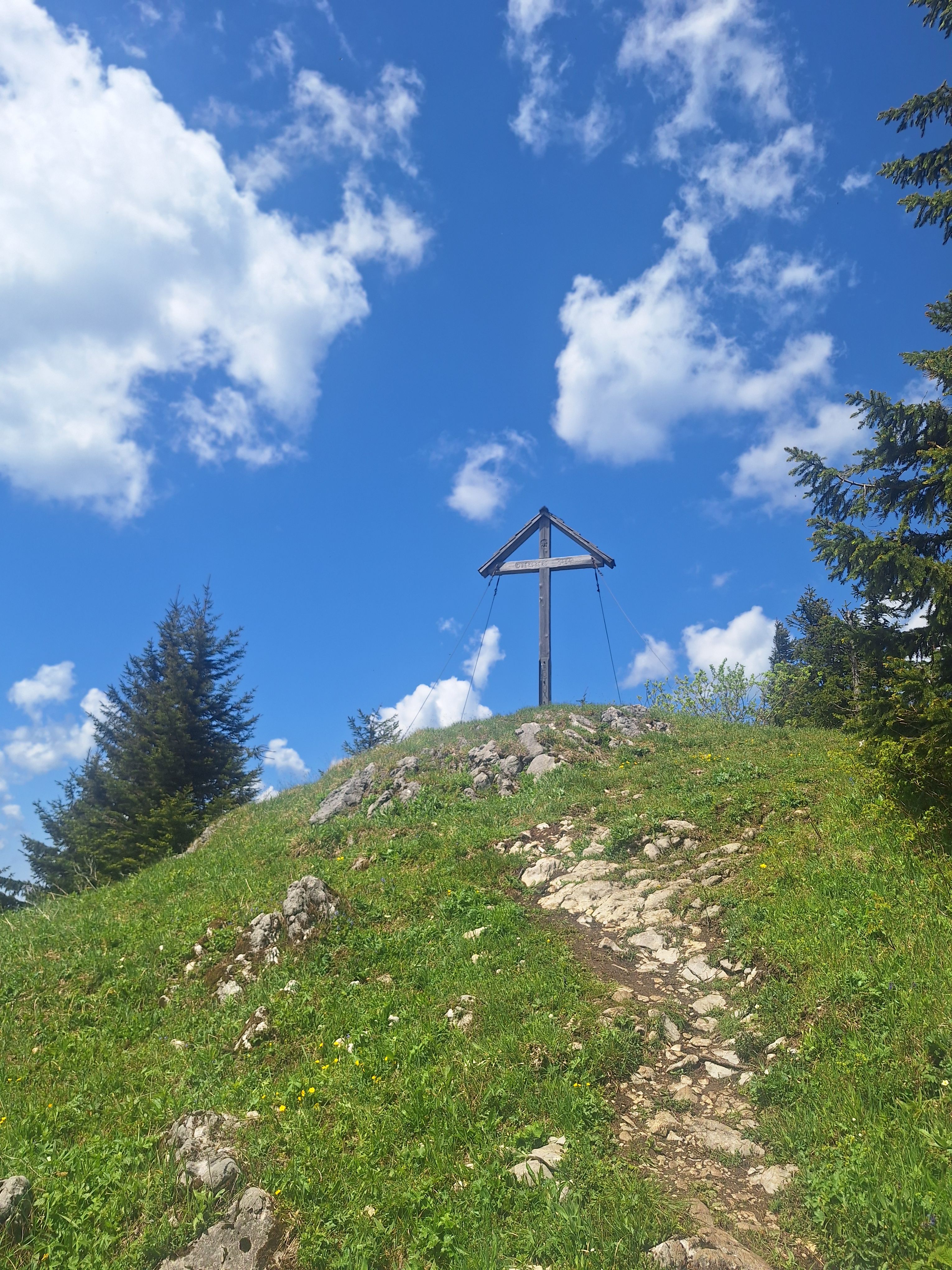 Gipfelkreuz auf einem Hügel mit blauem Himmel und Wolken.