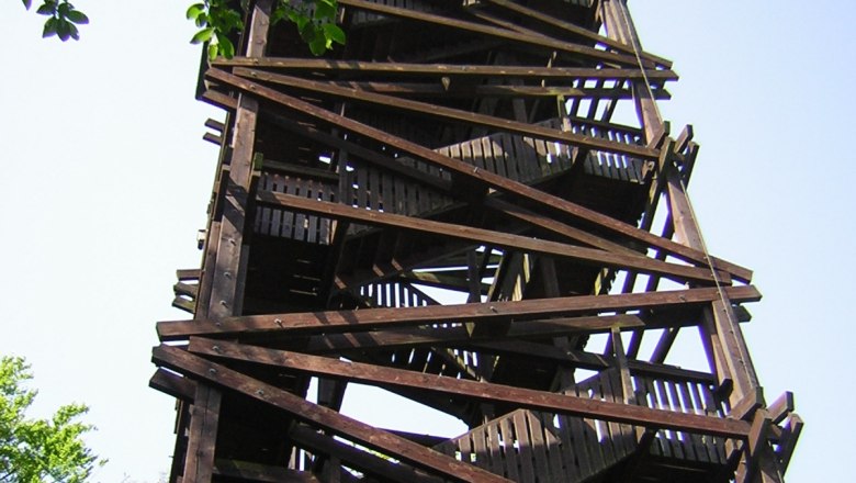 Holzturm mit Treppen und Geländern, umgeben von Bäumen.