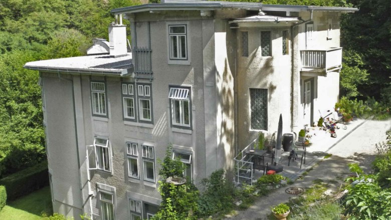 Großes, mehrstöckiges Haus mit grauer Fassade, umgeben von Bäumen und Garten.