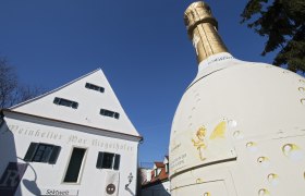 Gro&szlig;e Sektflasche vor einem wei&szlig;en Geb&auml;ude mit der Aufschrift 'Weinkeller Max Riegelhofer'.