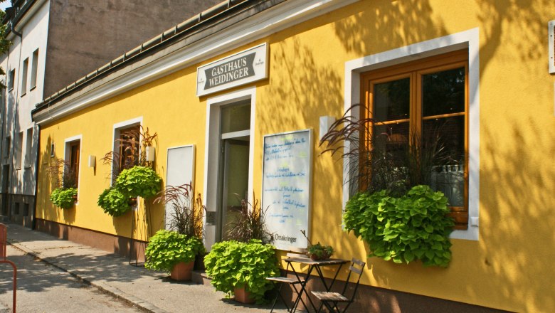 Gelbes Geb&auml;ude mit der Aufschrift 'Gasthaus Weidinger', Fenster mit Blumenk&auml;sten und ein Tisch mit Stuhl davor.