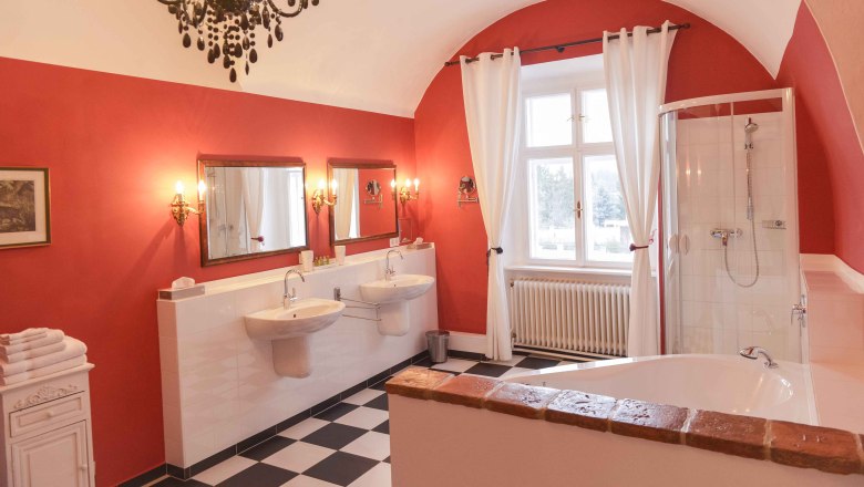 Elegantes Badezimmer mit roten Wänden, zwei Waschbecken, Badewanne, Dusche und Kronleuchter.