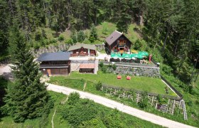 Luftaufnahme der Potschacherhütte in einem Waldgebiet mit umliegenden Bäumen und einer Terrasse mit Sonnenschirmen.