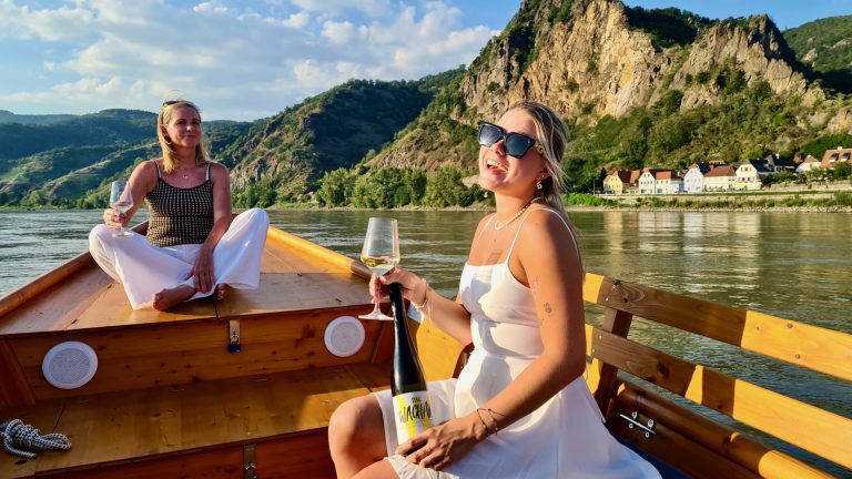 Zwei Frauen auf einem Boot in der Wachau, genießen Wein mit Blick auf die Berge.