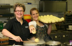 Zwei Frauen in einer K&uuml;che, eine h&auml;lt einen Tablett mit Kn&ouml;deln, die andere kocht Kn&ouml;del in einem Topf.