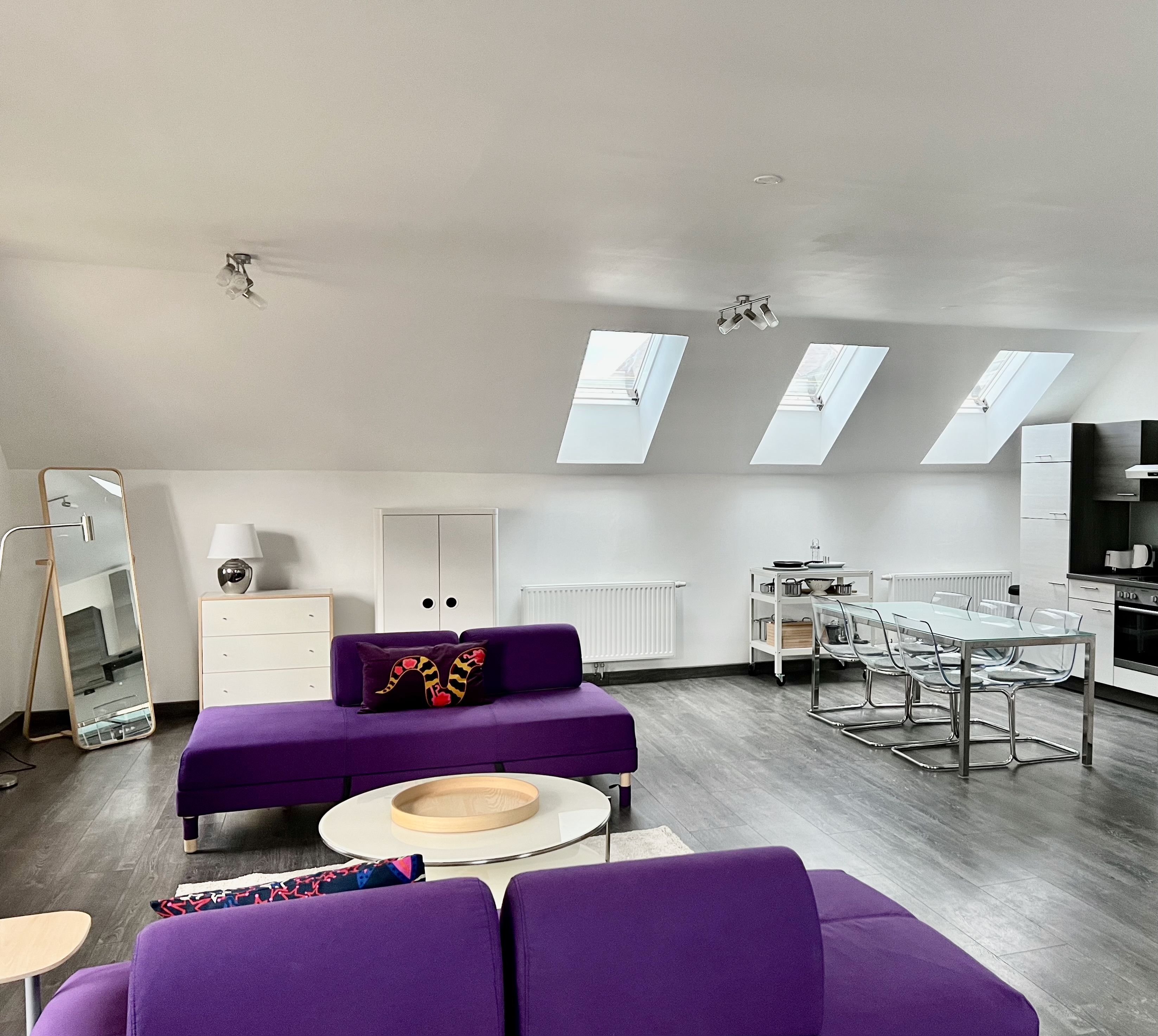 Modernes Loft mit violetten Sofas, Esstisch und Küchenzeile unter Dachfenstern.