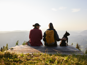 In der goldenen Abendsonne genie&szlig;en zwei Wanderer die atemberaubende Aussicht auf die sanften H&uuml;gel und das weite Tal. Ihr treuer Begleiter, ein kleiner Hund, sitzt entspannt neben ihnen und teilt diesen friedlichen Moment der Naturverbundenheit. Die frische Bergluft und die Stille der Umgebung laden dazu ein, die Seele baumeln zu lassen.