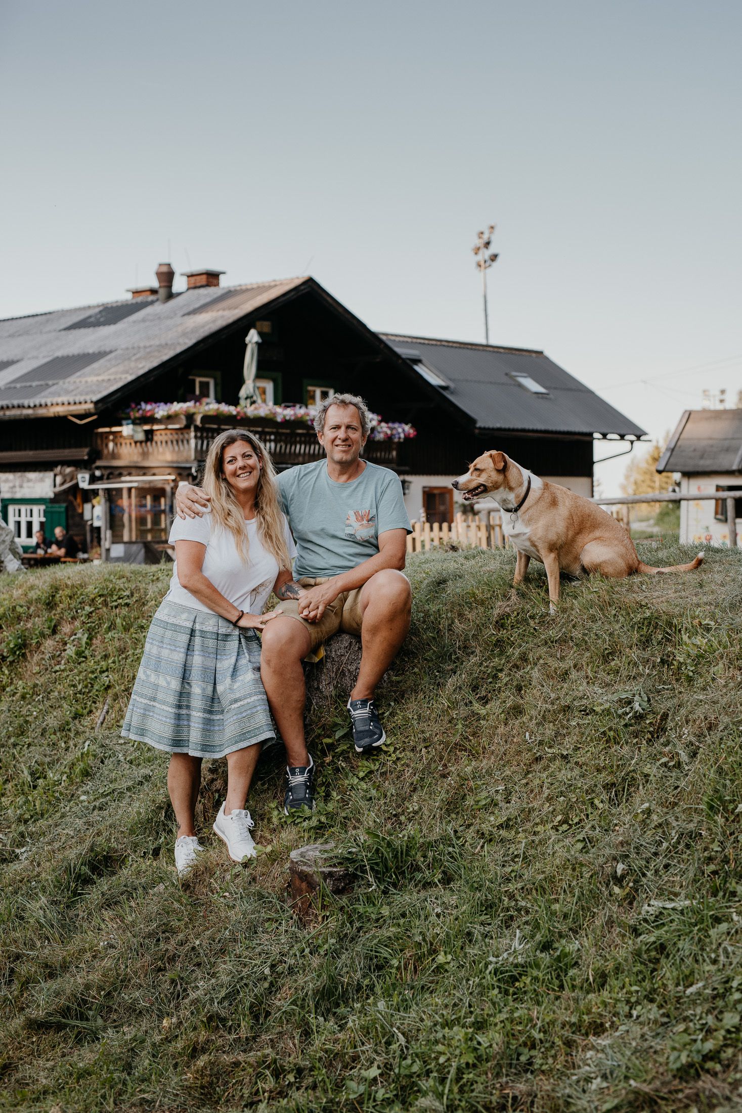 Ein Paar sitzt auf einem Hügel vor einer Hütte mit einem Hund daneben.