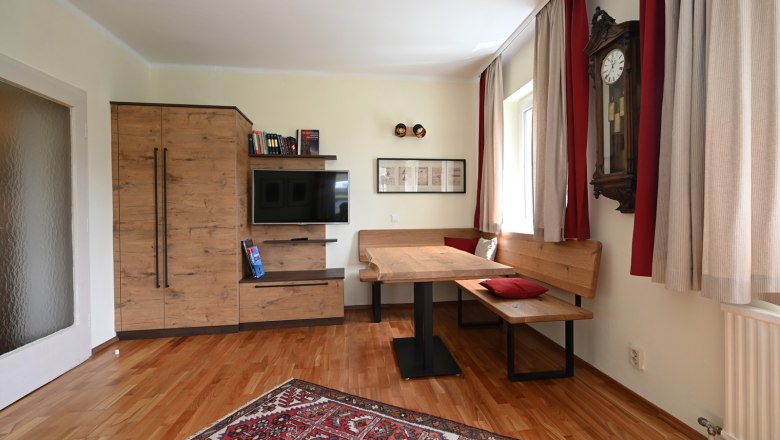 Gem&uuml;tliches Wohnzimmer mit Holzm&ouml;beln, einem Teppich, einer Eckbank und einer Wanduhr.