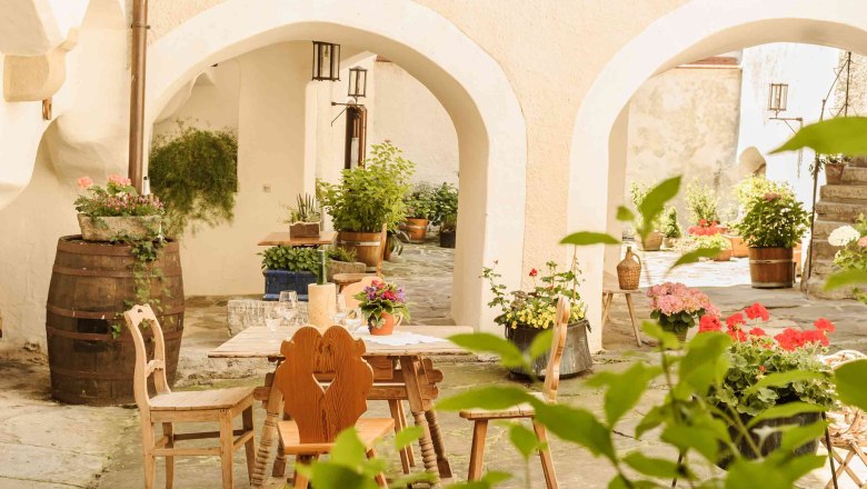 Ein gemütlicher Innenhof mit Holztisch und Stühlen, umgeben von Pflanzen und Blumen in Töpfen.