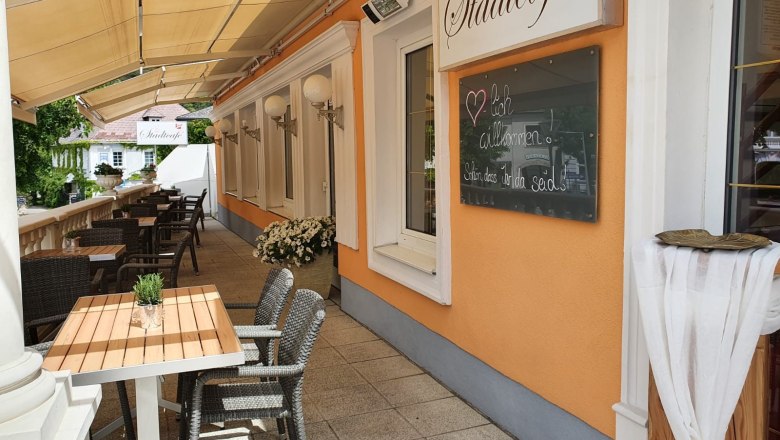 Au&szlig;enbereich eines Caf&eacute;s mit Tischen und St&uuml;hlen, orangefarbene Wand mit Schild 'Stadtcaf&eacute;'.
