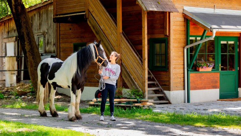 Eine Frau steht neben einem schwarz-wei&szlig;en Pferd vor einem Holzgeb&auml;ude.