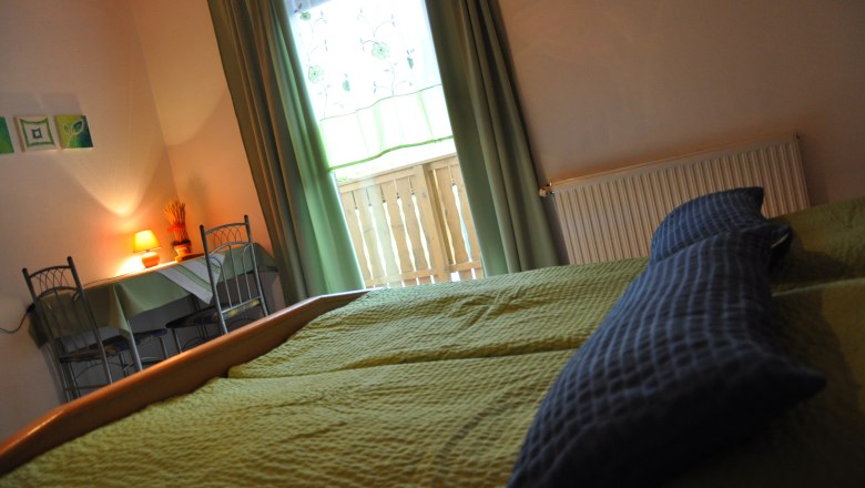 Gem&uuml;tliches Schlafzimmer mit gr&uuml;nem Bett, Tisch und St&uuml;hlen, Lampe und Fenster mit Vorh&auml;ngen.