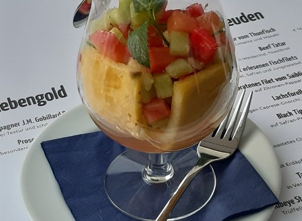 Ein Glas mit Fruchtsalat auf einem Teller mit blauer Serviette und Besteck.