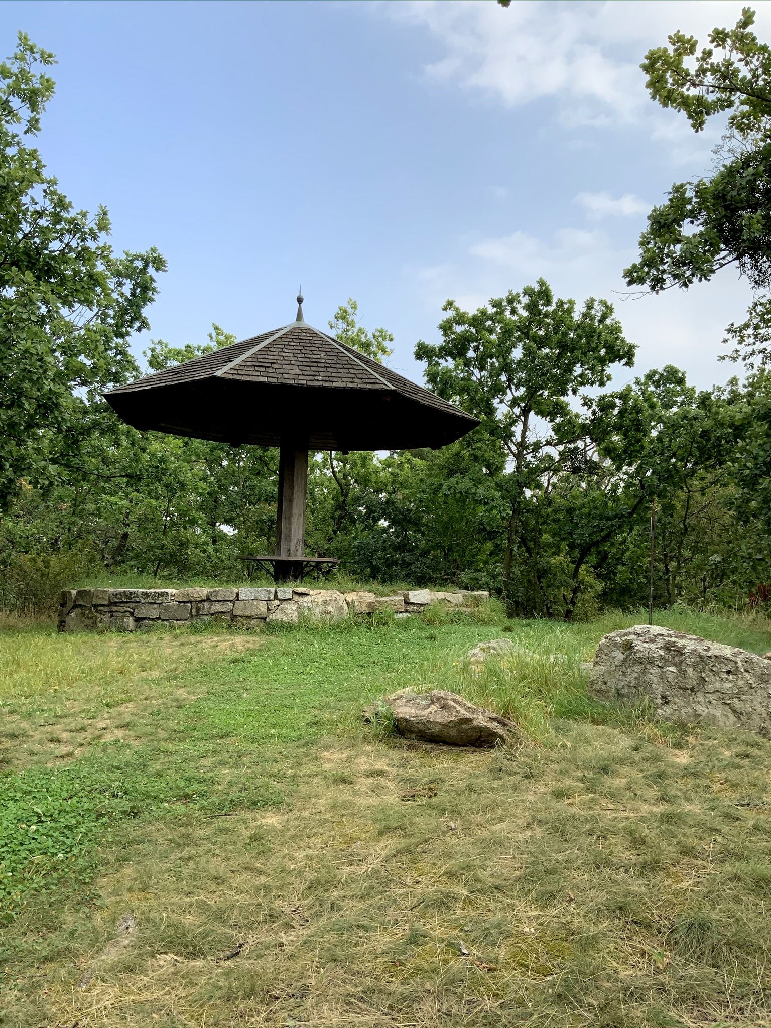 Ein hölzerner Pavillon mit spitzem Dach in einer grünen, bewaldeten Umgebung.