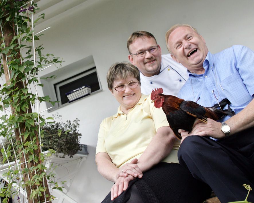 Drei lachende Personen sitzen auf einer Treppe, eine hält einen Hahn.