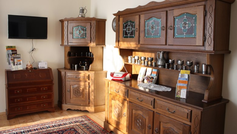 Wohnzimmer mit antiken Holzm&ouml;beln, einem Fernseher an der Wand und dekorativen Metallgef&auml;&szlig;en.