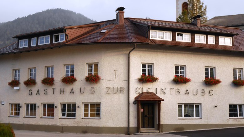 Hausansicht, © Gruber Johann Ein traditionelles Gasthaus mit Blumen an den Fenstern und einem Kirchturm im Hintergrund.