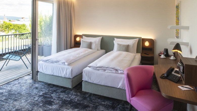 Modernes Hotelzimmer mit zwei Einzelbetten, einem Schreibtisch mit rosa Stuhl und Zugang zu einem Balkon mit Blick ins Grüne.