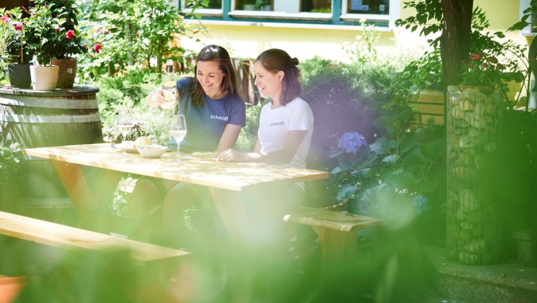Zwei Frauen sitzen an einem Holztisch im Garten und genießen Wein.