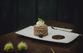 Ein Dessert aus Kastanienmousse auf einem weißen Teller, garniert mit Sahne und Minzblatt, daneben eine Schokoladensauce und eine Kirsche.