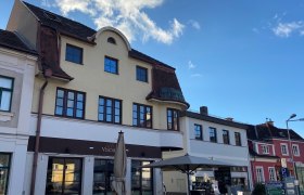 Außenansicht eines Cafés mit Terrasse und Sonnenschirm vor einem mehrstöckigen Gebäude.