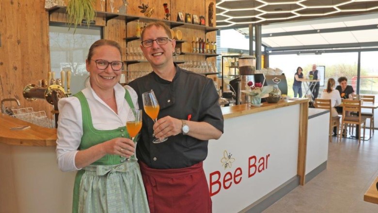 Zwei Personen in traditioneller Kleidung stehen lächelnd vor der Bee Bar mit Getränken in der Hand.
