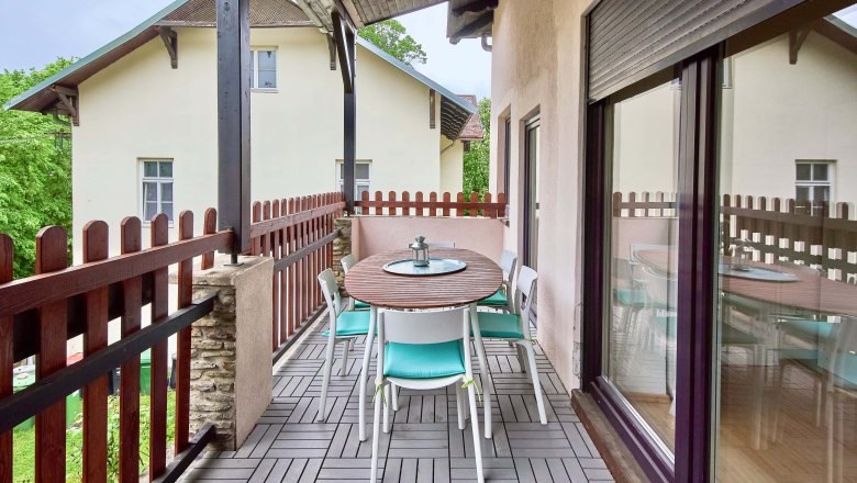 Ein Balkon mit Holztisch und Stühlen, umgeben von einem Holzzaun, mit Blick auf ein benachbartes Gebäude.