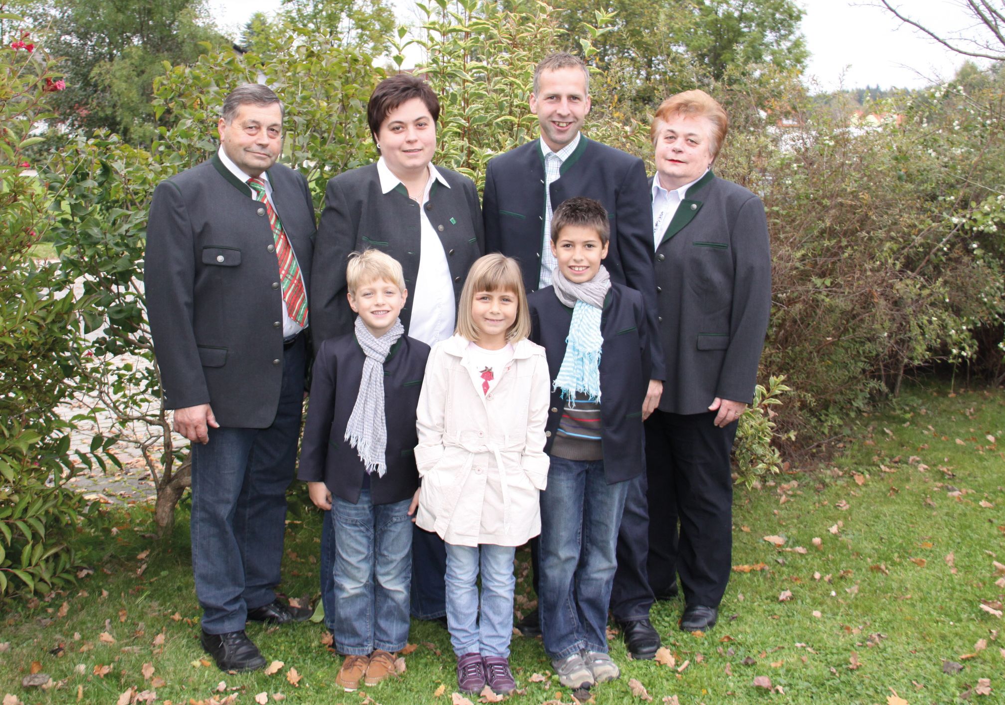 Eine Familie steht draußen in einem Garten, alle tragen formelle Kleidung. Im Hintergrund sind Büsche und Bäume zu sehen.