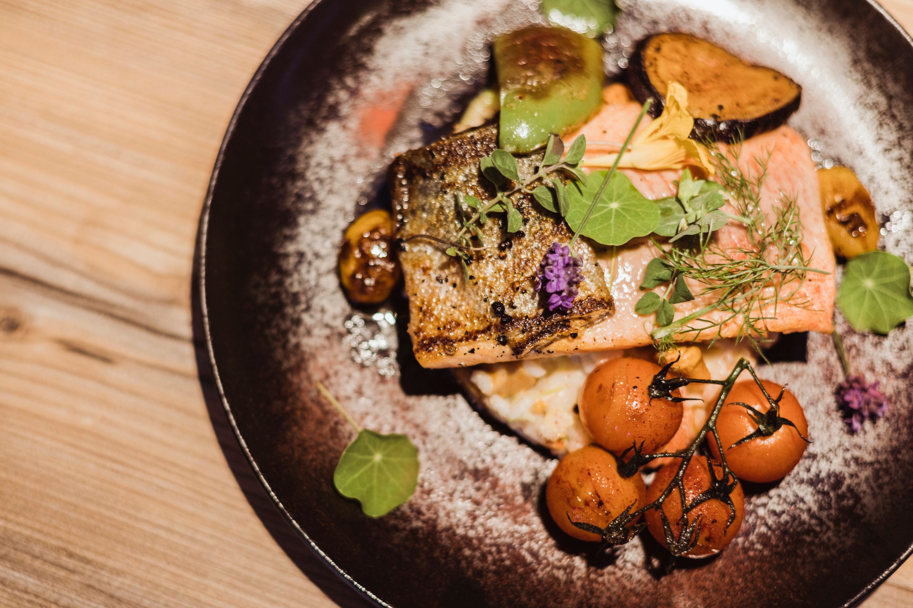 Lachsforellenfilet auf Grammelrisotto mit Gemüse und Kräutern auf einem Teller.