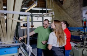 Drei Personen in einer Textilfabrik betrachten eine Maschine.