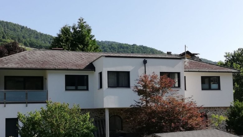 Außenansicht eines modernen weißen Ferienhauses mit Bäumen im Vordergrund und bewaldeten Hügeln im Hintergrund.