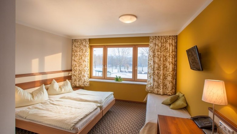 Ein gemütliches Doppelzimmer mit zwei Betten, gelben Wänden, einem Fenster mit Vorhängen, einem Fernseher an der Wand und einer Tischlampe.