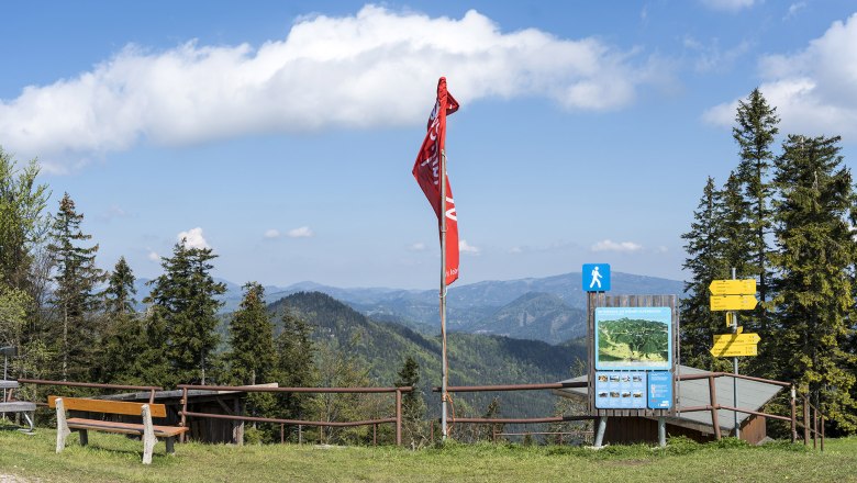 Aussichtspunkt mit Bank, roter Fahne und Wegweisern vor Berglandschaft.