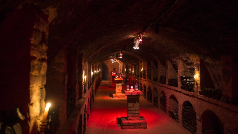 Ein stimmungsvoll beleuchteter Weinkeller mit Flaschen auf Steins&auml;ulen.