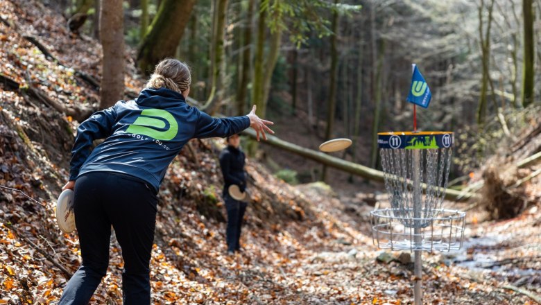 Person wirft Frisbee in Discgolf-Korb im Wald.