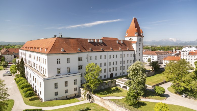 Blick auf eine historische Militärakademie mit rotem Dach und Turm, umgeben von grünen Bäumen und Rasenflächen, im Hintergrund Berge.