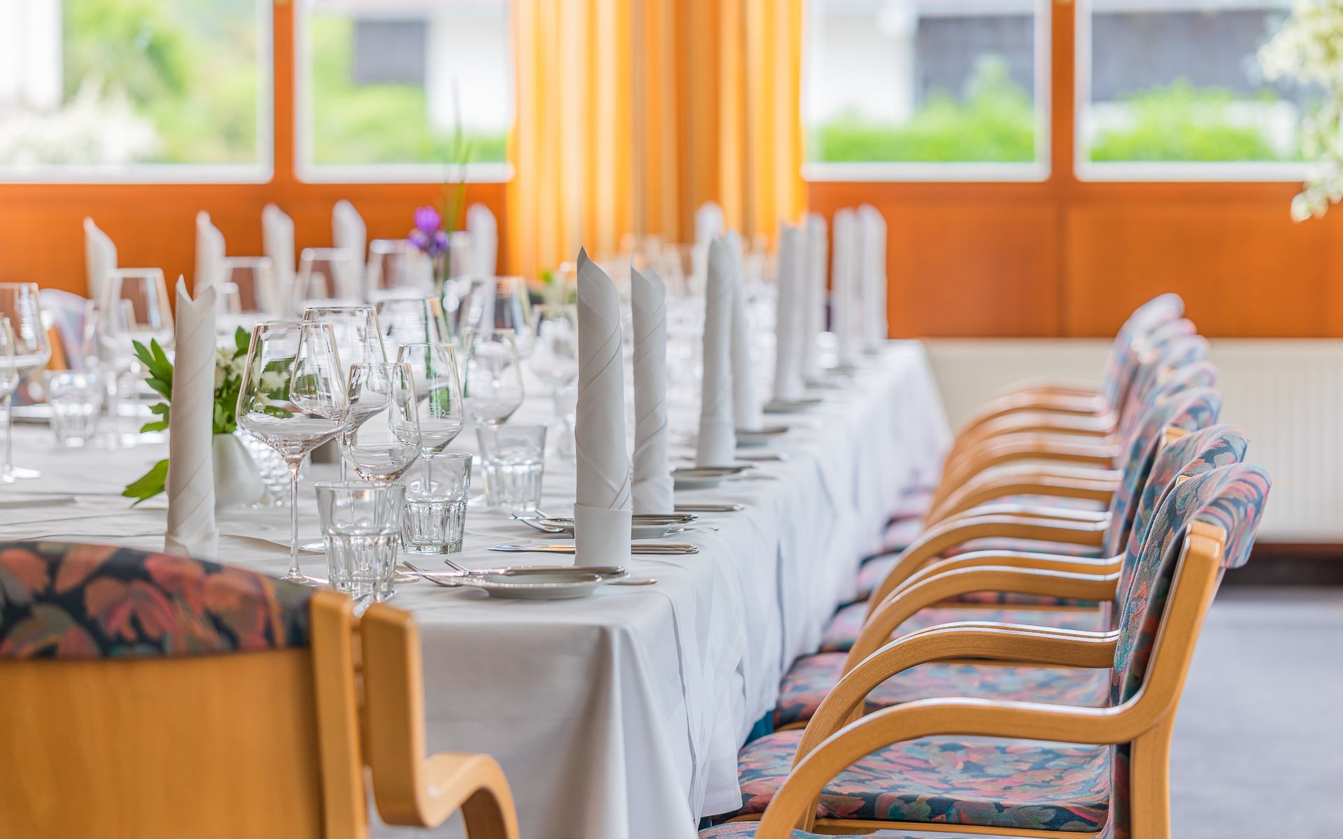 Elegant gedeckter Tisch in Wintergarten mit weißen Servietten, Gläsern und Besteck. Im Hintergrund große Fenster.